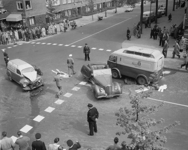 Schwarz-weiß-Szene eines Autounfalls an einer Straßenecke mit einem Van, anderen Fahrzeugen und einer Gruppe von Menschen in der Nähe, umgeben von Gebäuden, Bäumen, Masten und Schildern.