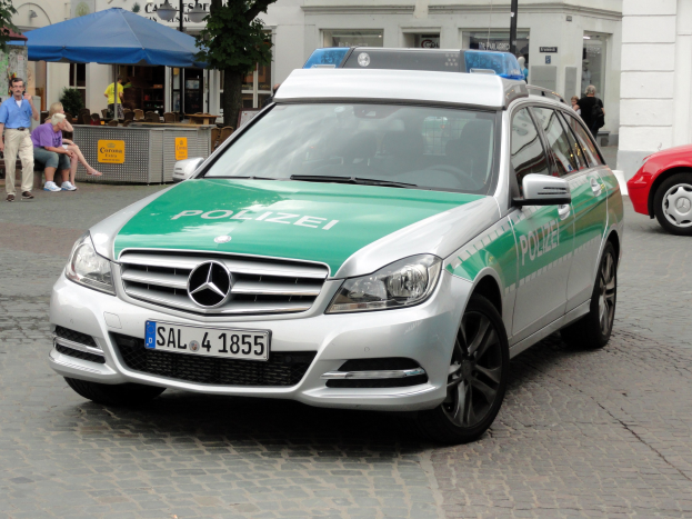 Polizeiauto neben einem roten Mercedes-Benz C-Klasse W204 auf einer Straße mit Gebäuden, Bäumen und Menschen im Hintergrund geparkt.