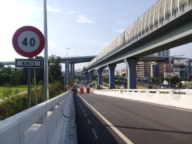 Autobahn mit Tempolimit-Schild, Fahrzeugen, Brücke, Laternenpfählen, Bäumen, Gebäuden und bewölktem Himmel im Hintergrund.