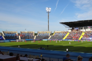 Ein Fußballspiel in einem großen, beleuchteten Stadion mit Zuschauern auf den Rängen und Spielern auf dem Feld unter einem sichtbaren Himmel.
