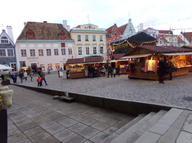 Ein belebter Weihnachtsmarkt in Tallinn, Estland mit Menschen um geschmützte Stände, festlicher Beleuchtung, bewölktem Himmel, Treppen führfühhrend und Topfpflanzen im Bereich.