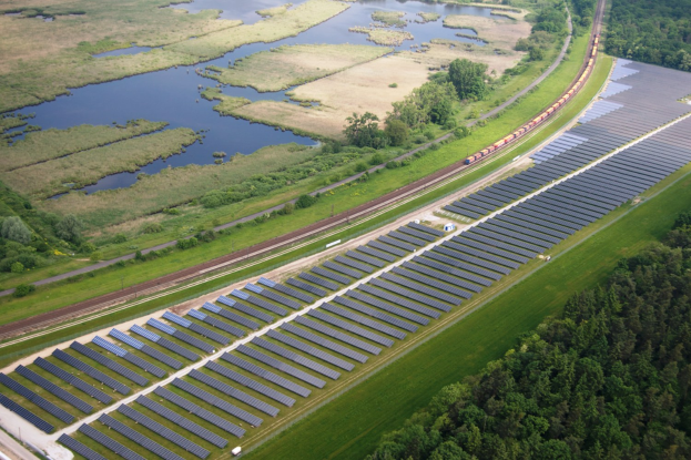 Luftaufnahme eines großen Solarparks auf einem Feld mit Bäumen, Gras, Pflanzen und Wasser, mit Solarpanelen und einem Zug auf einer Bahnstrecke.
