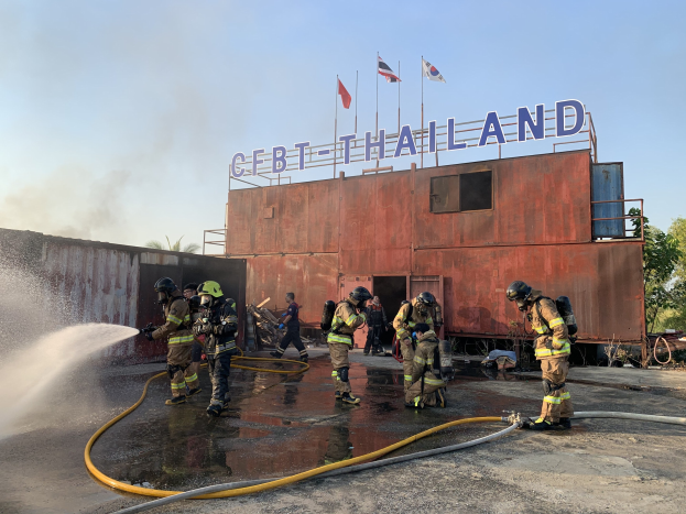 Feuerwehrmänner in Helmen sprühen Wasser auf ein Gebäude mit Text, während Fahnen, Bäume und ein klarer Himmel im Hintergrund zu sehen sind.