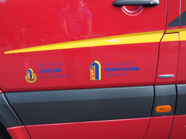 Ein roter Van mit einem gelben Streifen, einem Türgriff und einem Logo mit einem Löwen in einem Schild, umgeben von einem Lorbeerkranz, mit der Aufschrift "Deutsche Jugendfeuerwehr Verband".