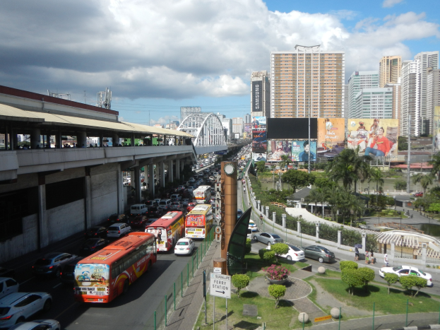 Eine belebte Stadtstraße mit hohen Gebäuden, Fahrzeugen, einer Brücke mit Fußgängern, Bäumen, Gras, Beschilderung und einem bewölkten Himmel.