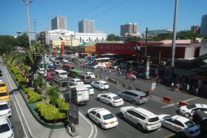 Eine belebte Stadtstraße mit Autos, Bussen, Fußgängern, Bäumen, Laternenmasten und Gebäuden unter einem klaren blauen Himmel.