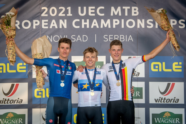 Drei medaillengewinnende Radrennfahrer stehen auf einem Podium und halten Blumensträuße in den Händen, im Hintergrund eine "2021 UEC MTB European Championships"-Fahne.