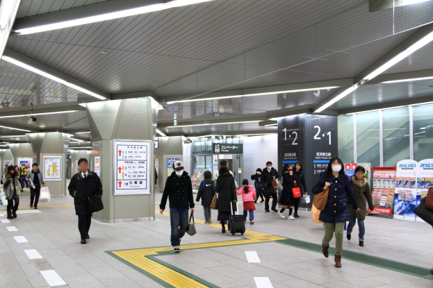 Menschen mit Gepäck durch einen Bahnhof mit Hinweistafeln und Deckenbeleuchtung.