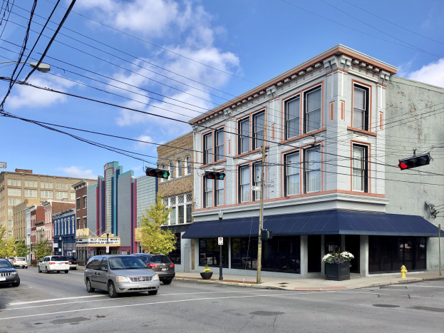 Eine Stra├če in einer Stadt mit parkenden Autos, Strommasten, Ampeln, Stra├čenlaternen, B├Ąumen und Geb├Ąuden mit Fenstern, unter einem bew├Âlktem Himmel, mit Text, der unten ein kommerzielles Grundst├╝ck zum Verkauf f├╝r $1,250,000 anzeigt.