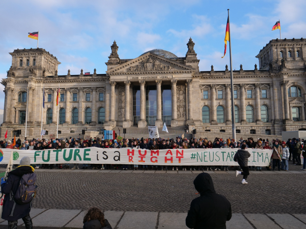 Gruppe von Menschen vor dem Reichstaggebäude in Berlin mit einem Banner mit der Aufschrift "Zukunft ist ein menschlicher Neustar ima", das Gebäude mit Fenstern, Säulen, Bögen und Statuen sowie umgeben von Fahnenmasten.