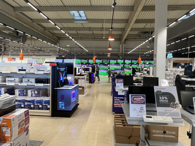 Ein großer Elektronikbereich in einem Walmart-Laden mit Regalen voller Computer, Drucker und Kartons, mit Menschen auf dem Boden, einer Säule, Schildern, Deckenlampen und Ventilatoren.