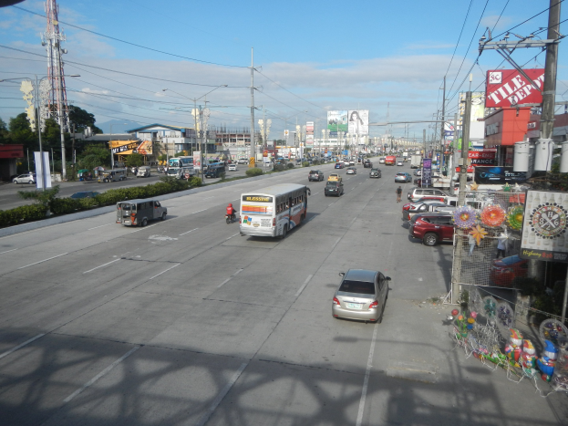 Eine belebte Stadtstraße mit verschiedenen Fahrzeugen, Gebäuden, Strommasten, Bäumen und einem Turm unter einem bewölkten Himmel.