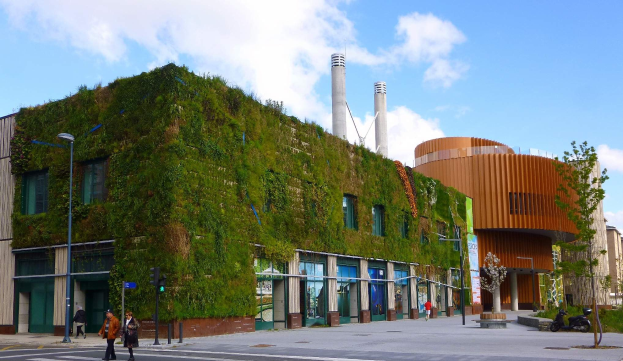Großes grünwandiges Gebäude mit Straßeninfrastruktur, Fahrzeugen, Fußgängern, Bäumen und bewölktem Himmel im Hintergrund.