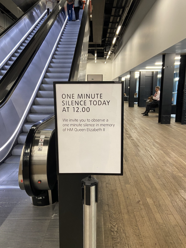 Eine Rolltreppe im Flughafen mit einem Schild, auf dem "Eine Minute Stille heute" steht, sowie ein paar Menschen darauf und an der Decke angebrachte Lichter im Hintergrund.
