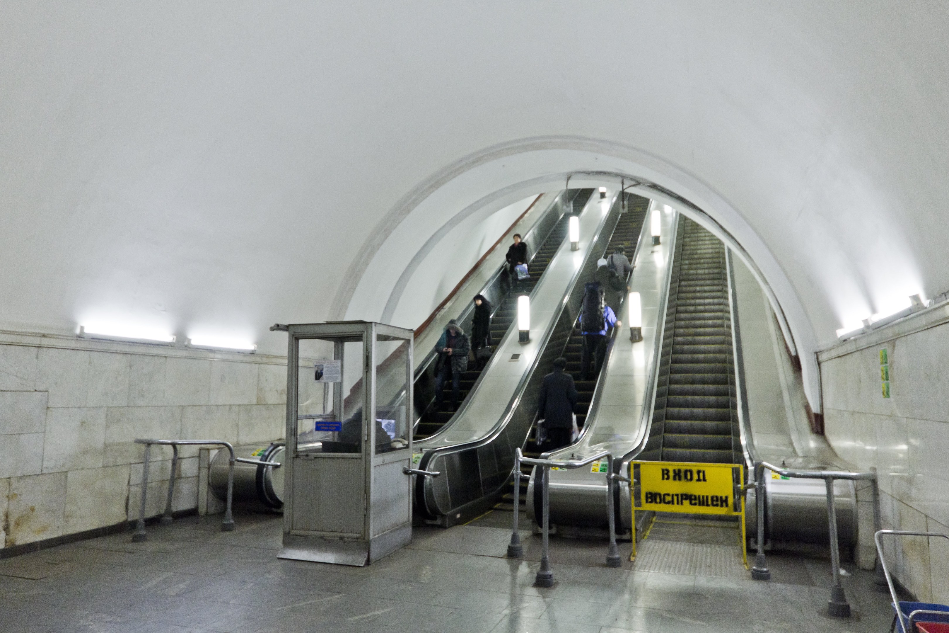 Rolltreppe in einer U-Bahn-Station mit Menschen, Geländern, Informationsschildern, Wandlampen und einem Einkaufswagen unten.