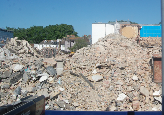 Großer Schutthaufen auf einer Baustelle mit einer Abrissmaschine unten, umgeben von Gebäuden, Bäumen und einem klaren blauen Himmel.