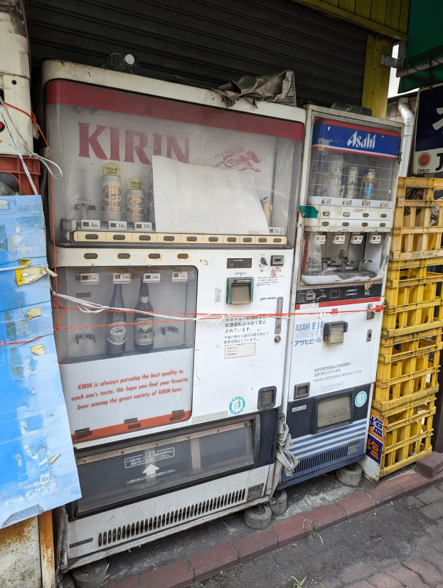 Ein Getränkeautomat auf einer Straße, gefüllt mit Flaschen, mit einem Rolladengebäude und einer Tafel mit Text im Hintergrund.
