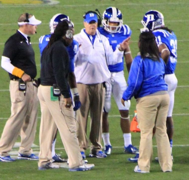 Gruppe von Menschen in Diskussion auf einem Fuballfeld, einige tragen Kappen und Helme, mit einer Wand im Hintergrund.