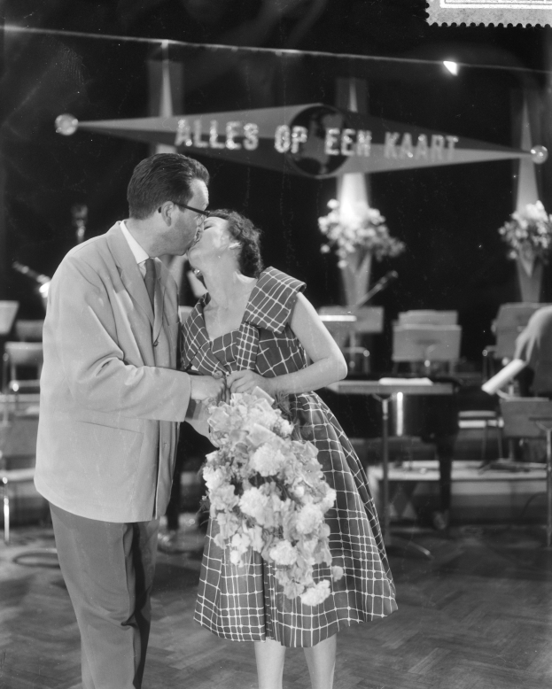 Ein Mann und eine Frau umarmen und küssen sich auf einer Bühne, wobei die Frau einen Blumenstrauß hält, umgeben von Stühlen, Blumenvasen und anderen Gegenständen.