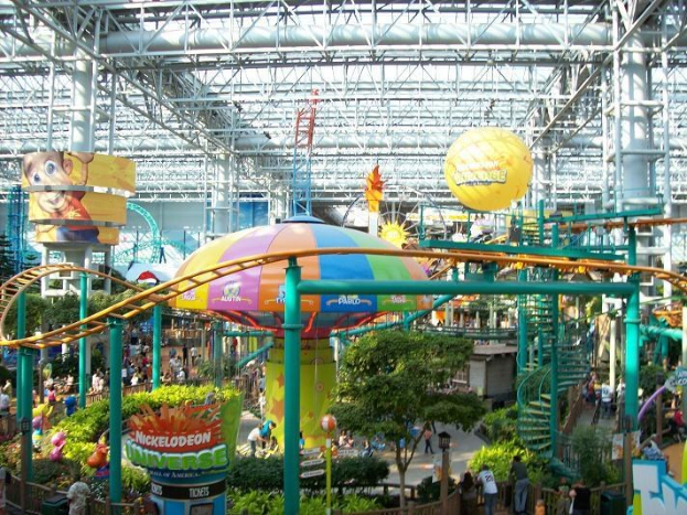 Indoor-Wasserpark mit einer Achterbahn umgeben von Menschen, Bäumen, Pflanzen und Deckenleuchten mit Stangen.