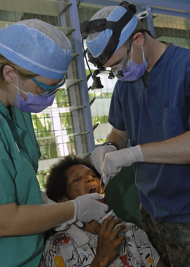 Zwei maskierte und handschuhgetragene Chirurgen führen eine Gesichtsoperation an einer Frau durch, die auf einem Bett liegt, mit Bäumen im Hintergrund.