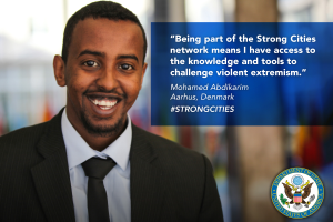 Ein Mann im Anzug und Krawatte lächelt in die Kamera mit einem Logo und Text auf der rechten Seite, der besagt "Teil des starken Städtenetzwerks zu sein bedeutet, Zugang zu Wissen und Werkzeugen zu haben, um gewalttätigen Extremismus infrage zu stellen", vor einem unscharfen Hintergrund.