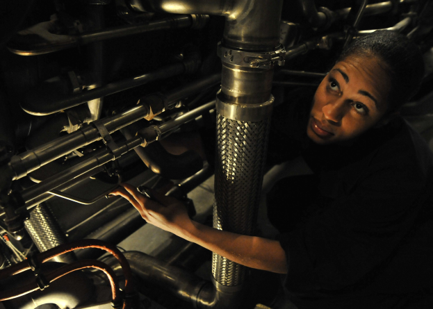 Ein Techniker in Uniform arbeitet konzentriert an einer Maschine in einem schwach beleuchteten Raum voller Rohre und Geräte.