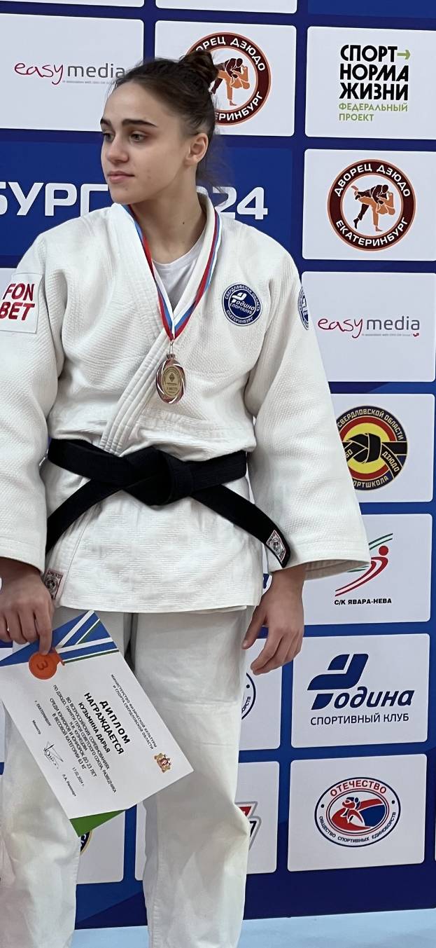 Frau in weißem Judo-Anzug mit schwarzem Gürtel auf einem Podium stehend, eine Medaille und ein Zertifikat haltend, mit einem Logo-Board im Hintergrund.
