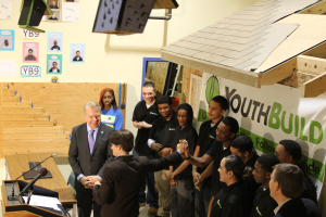 Eine Gruppe von Menschen steht um ein Rednerpult herum vor einer Menge, einige schütteln Hände, ein Tisch mit Mikrofonen und Papieren links, Plakate an der Wand im Hintergrund und Gegenstände auf dem Dach und der rechten Seite des Bildes.