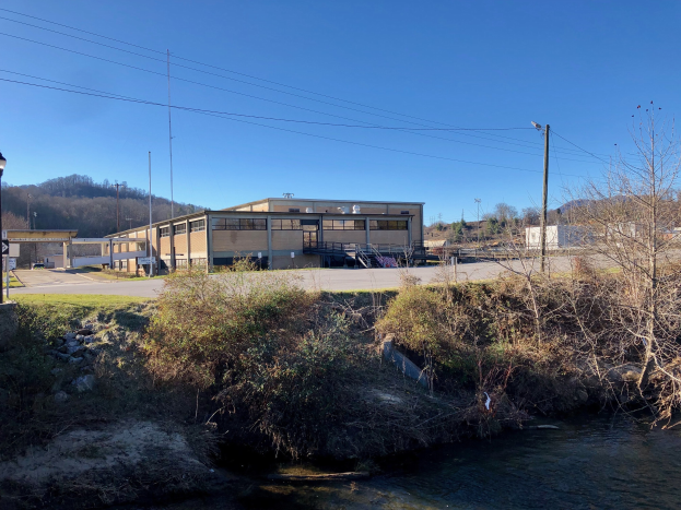 Ein Fluss fließt durch ein grünes Feld neben einem Gebäude in der Nähe von Bäumen, Pflanzen, Gras, Steinen, Straßenlaternen, Strommasten, Stromkabeln, Hügeln und einem klaren blauen Himmel, wobei das Gebäude als Standort einer Wasseraufbereitungsanlage geplant ist.