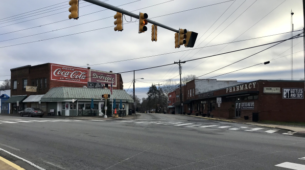 Eine Stadtstraßenkreuzung mit Ampel, Fahrzeugen, Gebäuden mit Schildern, Strommasten und -leitungen, Bäumen und einem bewölkten Himmel.