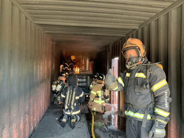 Feuerwehrleute in Schutzausrütstücken innerhalb eines Seecontainers, einer hält ein Rohr, mit einem Feuerlöscher im Hintergrund sichtbar.