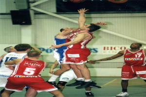 Eine Gruppe von Männern, die Basketball auf einem Court spielen, mit einer Wand im Hintergrund, die Text und Lautsprecher zeigt, und einem Basketball in der Luft oben auf dem Bild.