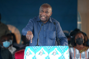 Ein Mann in einem blauen Hemd spricht in zwei Mikrofone an einem Rednerpult, während im Hintergrund eine Menge und mehrere sitzende Personen zu sehen sind, von denen einige Masken tragen.