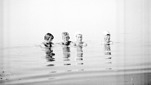Vier Kinder in Schwarz-Weiß schwimmen in einem Pool, jeder trägt eine Schwimmkappe.