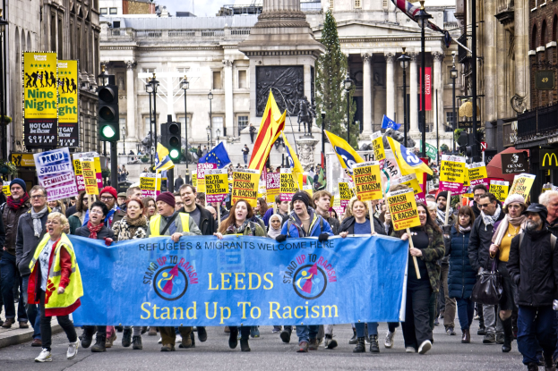 Eine Gruppe von Menschen marschiert mit einem 'Steh Auf gegen Rassismus'-Schild die Straße entlang, mit Gebäuden, Bäumen, Laternen, Verkehrszeichen und einer Statue im Hintergrund bei bewölktem Himmel.