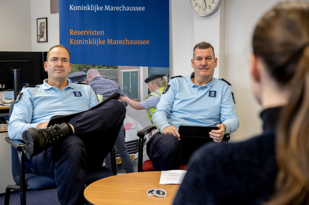 Zwei Polizeibeamte sitzen an einem Tisch mit Papieren und Gegenständen und sprechen mit einer Frau, mit einer Fahne, Uhr und Fotorahmen an der Wand und einem weiteren Tisch mit einem Monitor, Tastatur und anderen Gegenständen links daneben.