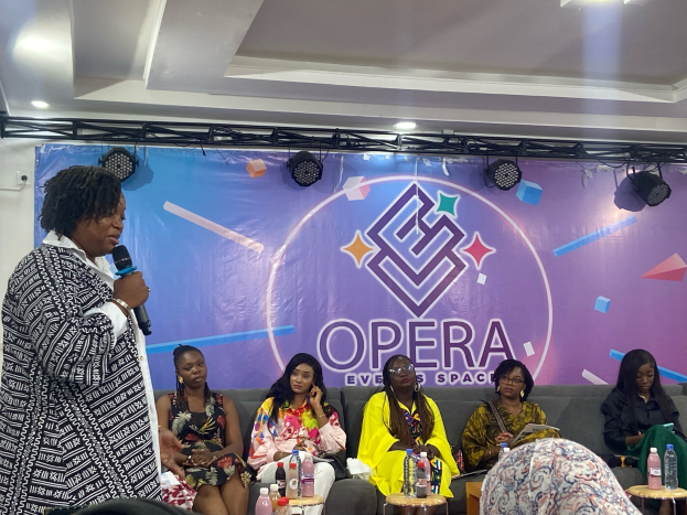 Frau mit Mikrofon steht vor einer Gruppe sitzender Frauen auf einem Sofa, mit Flaschen und Gegenständen auf den Tischen im Vordergrund und einem Banner mit Text und Deckenbeleuchtung im Hintergrund.