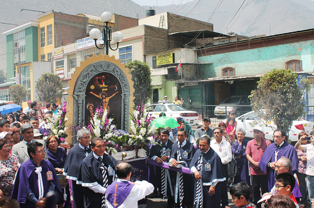 Menschen auf einer Straße außerhalb der Stadt, mit einem Kreuz und Blumensträußen im Vordergrund, fahrende Autos im Mittelgrund und Gebäuden, Bäumen und Straßenlaternen im Hintergrund.