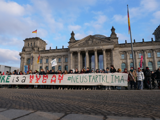 Gruppe von Menschen mit einem Banner mit der Aufschrift "Wir sind ein Menschenrecht" vor dem Reichstagsgebäude in Berlin, Deutschland, mit seinen architektonischen Details und umgeben von Fahnen unter einem bewölkten Himmel.