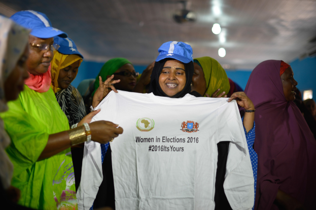 Eine Gruppe von Frauen, die zusammenstehen und ein weißes T-Shirt mit der Aufschrift 'Frauen in Wahlen 2016' hochhalten, einige tragen blaue Kappen, mit Beleuchtung im Hintergrund.