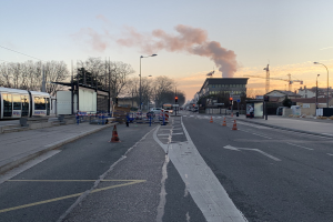 Straßenszene mit Absperrgittern, einem Bus, Laternen, Bäumen, Gebäuden, Kränen und Rauch, der aus einem entfernten Gebäude aufsteigt.