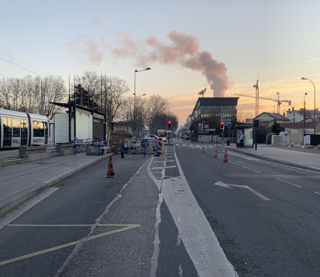 Straßenszene mit Absperrgittern, einem Bus, Laternen, Bäumen, Gebäuden, Kränen und Rauch, der aus einem entfernten Gebäude aufsteigt.
