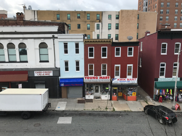 Luftaufnahme einer belebten Straße in Brooklyn, New York, mit Fahrzeugen, kommerziellen Geschäftsfronten, Werbetafeln, Strommasten und Drähten vor einem bewölkten Himmel.