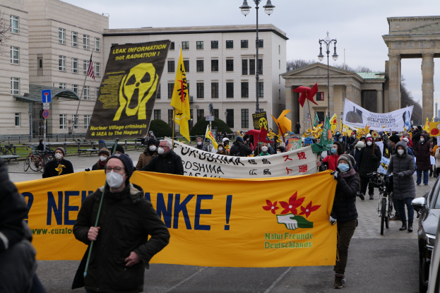 Große Gruppe von Menschen bei einer Straßendemonstration gegen Atomkraft in Deutschland, mit Schildern und Fahnen, Fahrzeugen auf der rechten Seite, Gebäuden und Bäumen im Hintergrund und einem Bogen weiter unten auf der Straße.