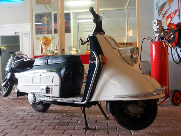 Ein weißes und schwarzes Vespa-Fahrrad, das vor einer Tankstelle geparkt ist, mit einem roten Feuerlöscher rechts daneben und einer Person links daneben, vor einer Glaswand mit Plakaten im Hintergrund.