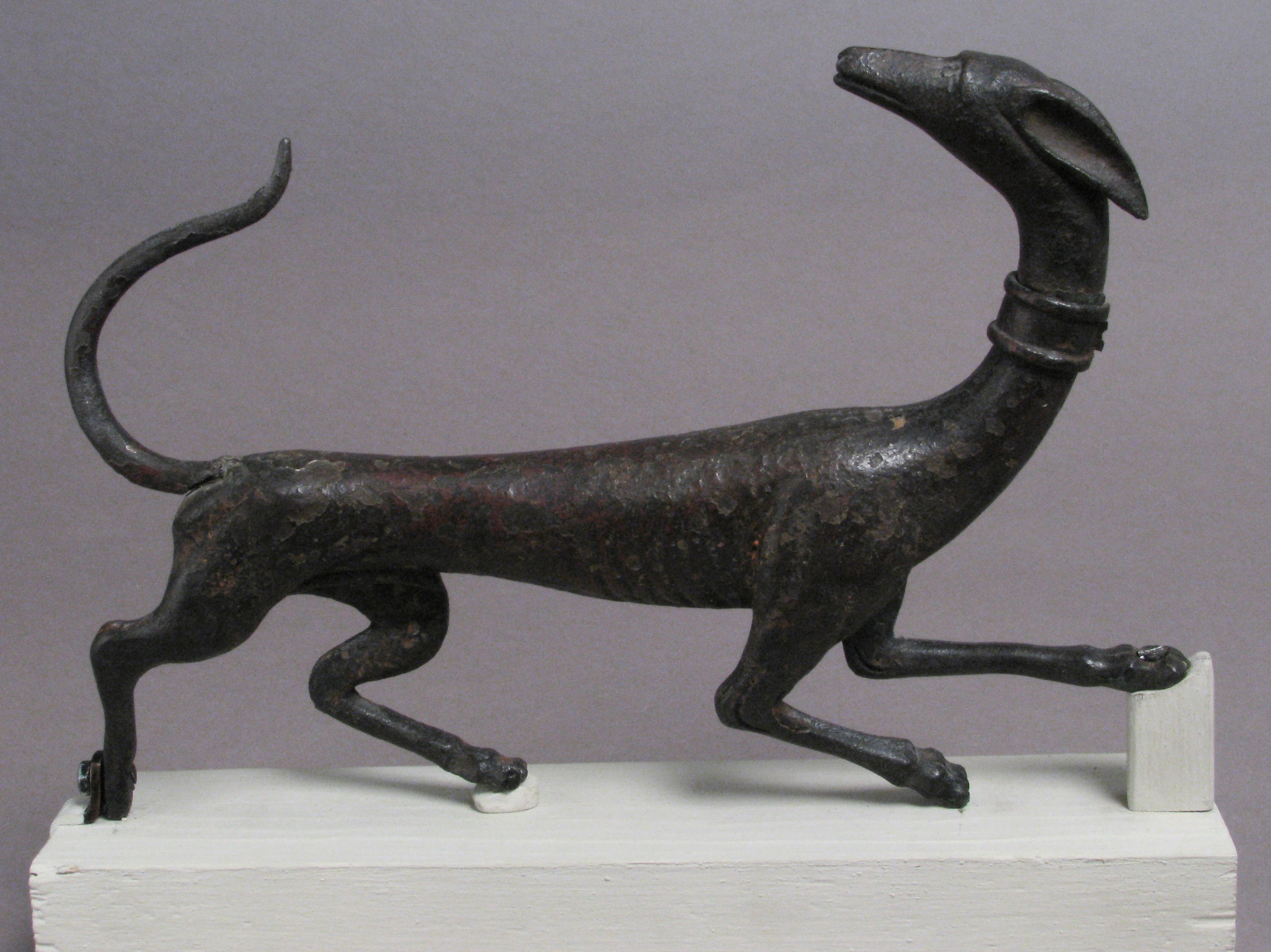 Eine Bronzestatue eines Greyhounds auf einem weißen Sockel vor einem grauen Hintergrund, mit detaillierten Darstellungen des Fells und der Merkmale des Hundes.