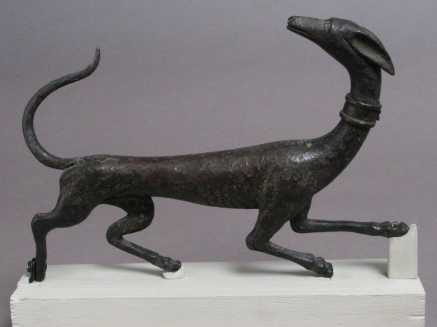 Eine Bronzestatue eines Greyhounds auf einem weißen Sockel vor einem grauen Hintergrund, mit detaillierten Darstellungen des Fells und der Merkmale des Hundes.