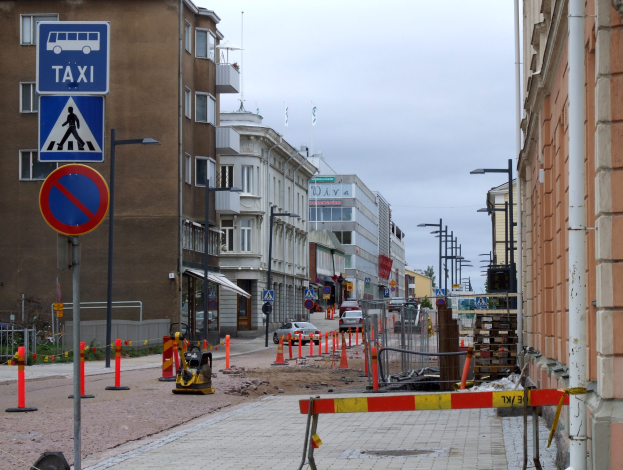 Eine Stadtstraße mit Gebäuden, Straßenlaternen, Schildern, Verkehrsleitkegeln, Fahrzeugen, Absperrpoller, Bäumen, einer Baustelle mit mehreren Verkehrsschildern und einem bewölkten Himmel.