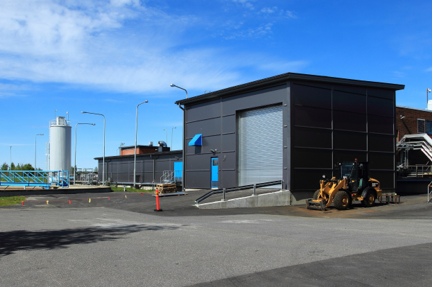 Großes Gebäude mit einem Bagger davor, umgeben von Straßeninfrastruktur, Fahrzeugen, Bäumen und einem bewölkten Himmel, als das neue Gebäude am Gewerbepark bezeichnet.